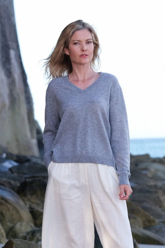 Woman on coastal rocks in grey cashmere V-neck and white pants – Love Cashmere NZ